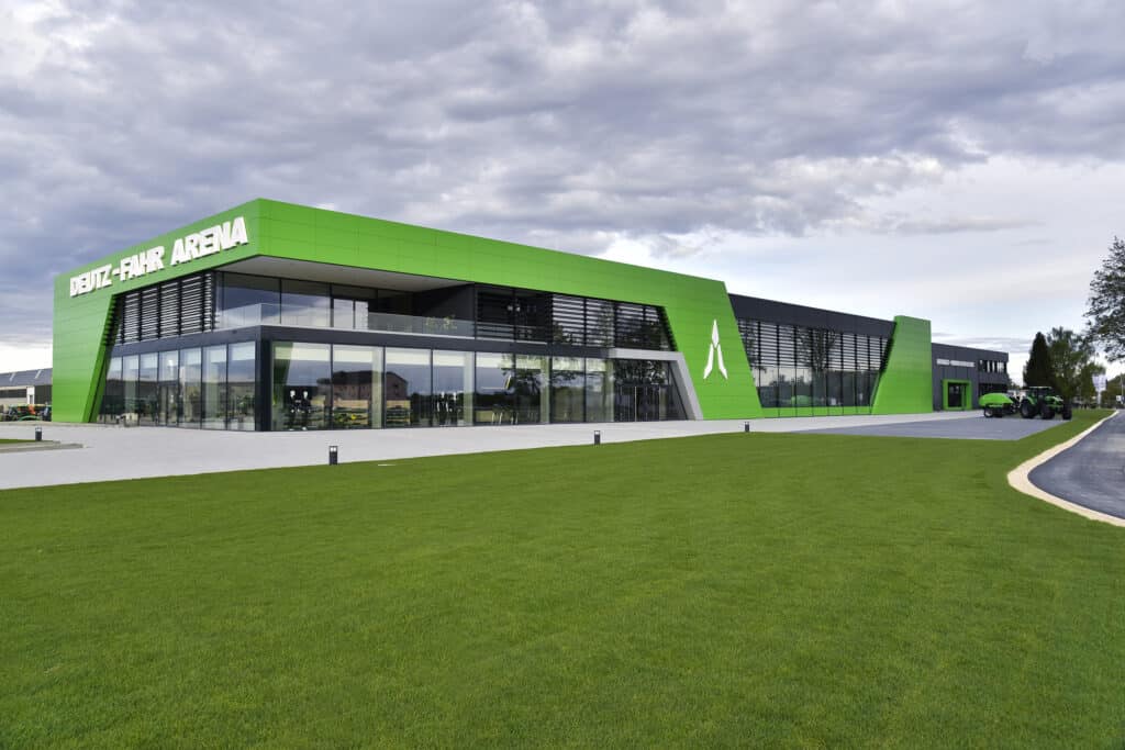 Moderne, weitläufige Ausstellungs- und Museumsanlage in markantem Deutz-Fahr-Grün. Die Fassade besteht aus großen Glasflächen und geometrischen, grün verkleideten Elementen. Vor dem Gebäude erstreckt sich eine große Rasenfläche, rechts stehen mehrere landwirtschaftliche Maschinen.