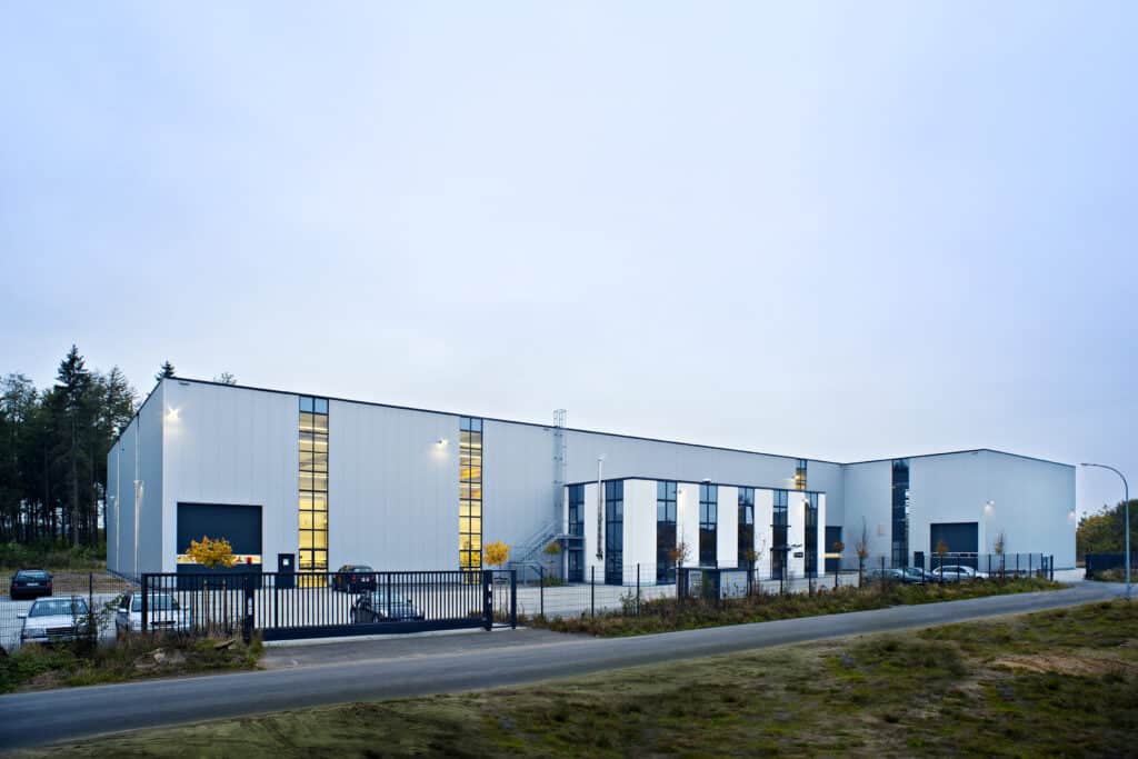 Neubau einer modernen Kranbahnhalle mit angrenzenden Büroanbauten in Stade bei Hamburg. Die großvolumige Industriehalle besitzt eine helle Metallfassade mit hohen vertikalen Fensterbändern, die den Innenraum großzügig belichten. Vor der Halle befindet sich ein umzäunter Betriebsbereich mit Parkplätzen, Toranlage und Außentreppen. Die klare, funktionale Architektur verbindet leistungsfähige Hallentechnik mit zeitgemäßen Bürostrukturen für Produktion und Verwaltung.