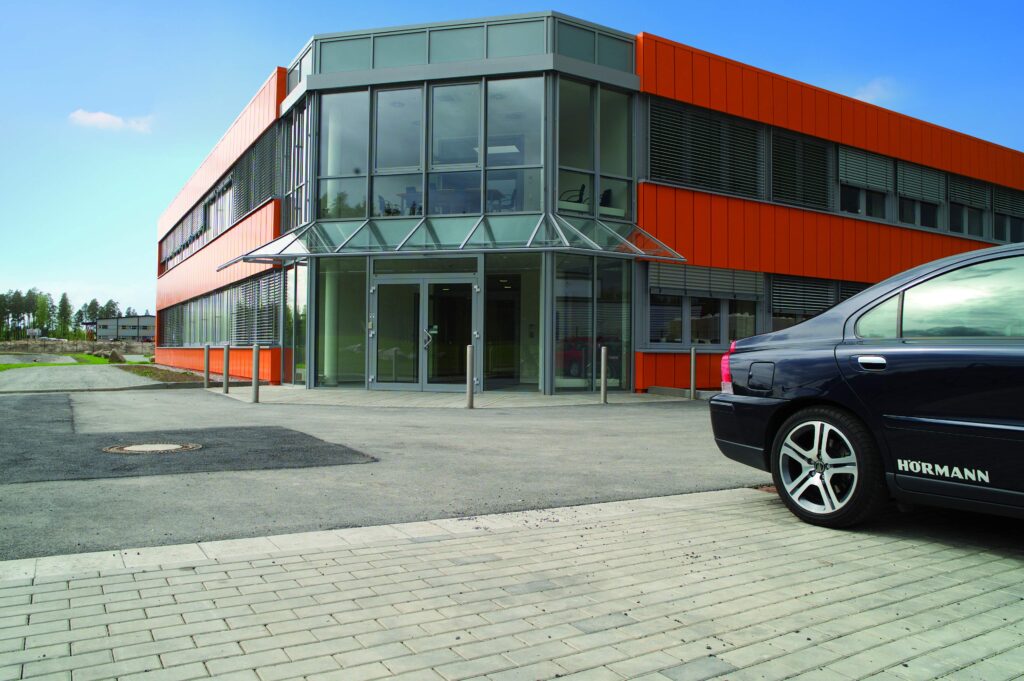 Modernes Büro- und Verwaltungsgebäude mit orangefarbener Fassade und Glas-Eingangsbereich, davor ein Parkplatz mit einem Auto.