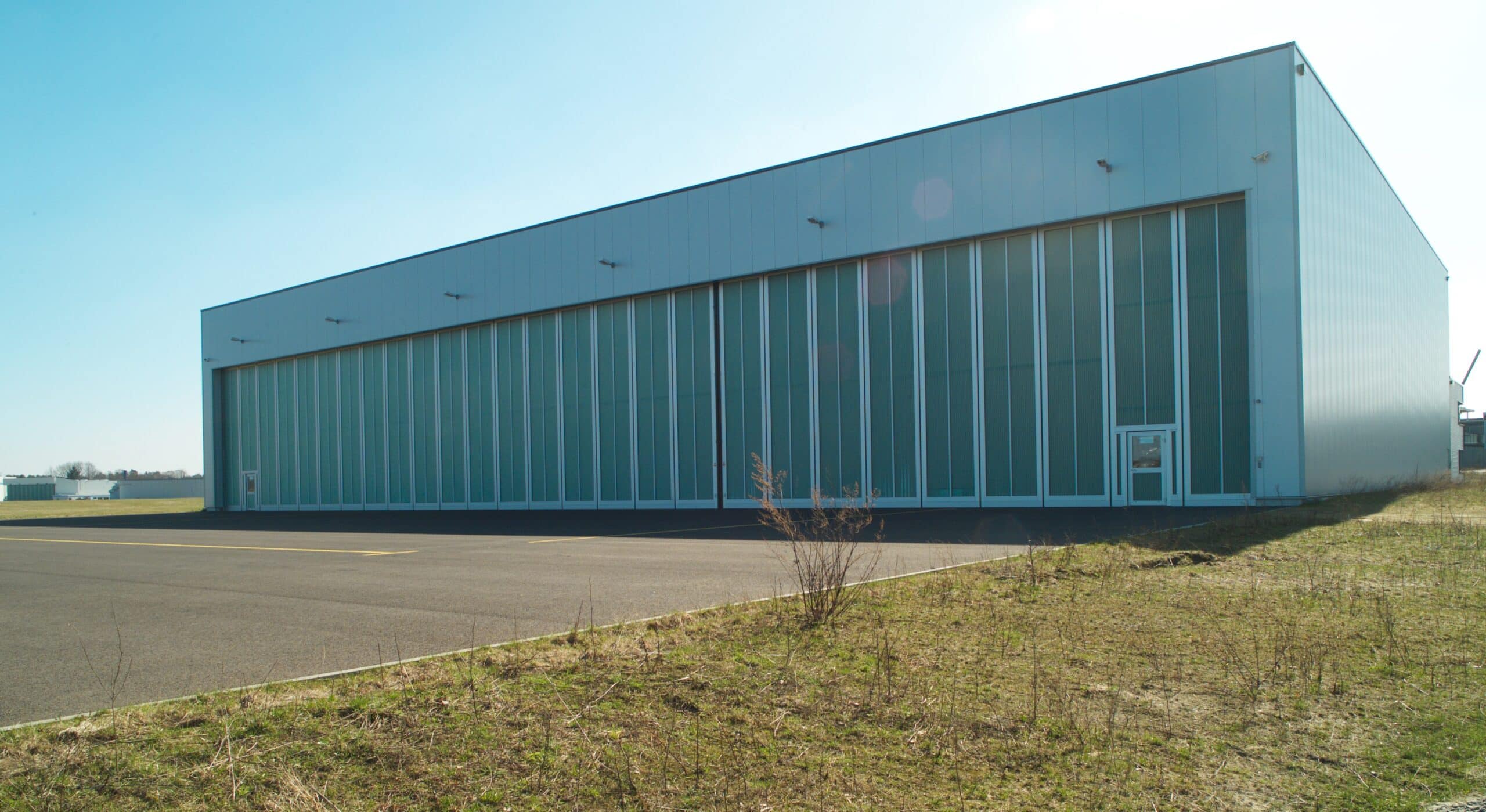 Außenansicht eines modernen Flugzeughangars in Braunschweig. Das große Gebäude mit lichtdurchlässigen Fassadenelementen und breiten Schiebetoren steht direkt am Rollfeld und bietet viel Platz für die Unterbringung und Wartung von Flugzeugen.