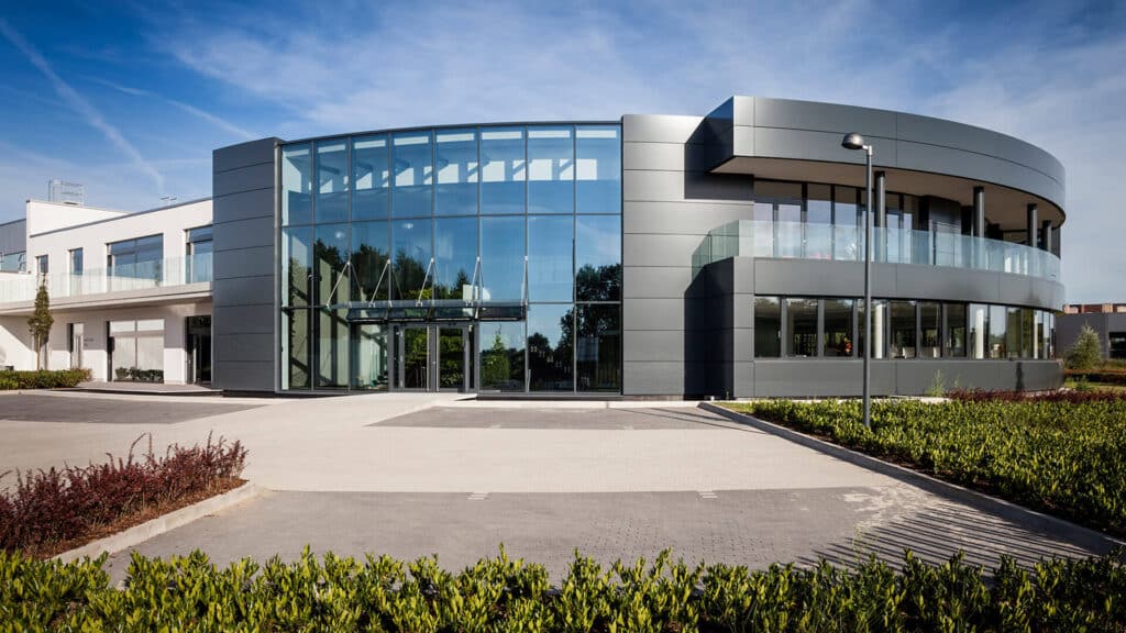 Modernes Verwaltungsgebäude mit großer, halbkreisförmiger Glasfassade im Eingangsbereich. Die Fassade kombiniert dunkle Metallverkleidung mit großflächigen Fensterfronten. Rechts befindet sich ein abgerundeter Gebäudeteil mit verglastem Balkon. Im Vordergrund sind gepflegte Grünanlagen und ein breiter, gepflasterter Vorplatz zu sehen.