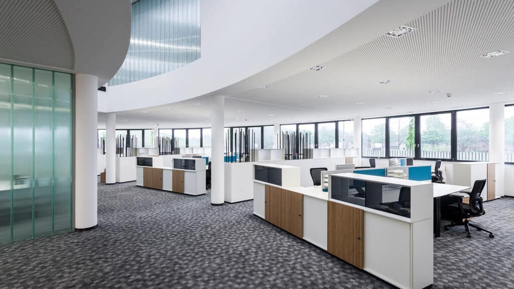 Großzügiges, modernes Großraumbüro mit hellen Arbeitsinseln, weißen Trennmöbeln und Holzakzenten. Große Fensterfronten lassen viel Tageslicht hinein. Die Decke ist glatt und hell, getragen von weißen Rundsäulen. Der Boden ist mit grauem, gemustertem Teppich ausgelegt. Links befindet sich eine halbtransparente Glaswand, darüber eine offene Galerie mit transluzenten Elementen.