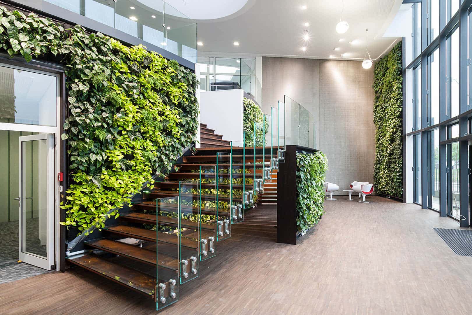 Großzügiges, modernes Foyer mit bodentiefen Glasfassaden, einer frei schwebenden Holztreppe mit Glasgeländer und üppigen vertikalen Begrünungen an mehreren Wänden.