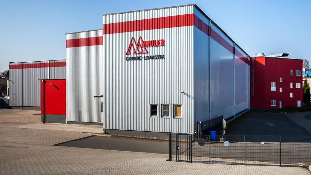 Sanierte Lagerhalle mit Verladebereich in Remscheid: Großes Industriegebäude mit grauen, vertikal profilierten Metallfassaden und roten Akzentflächen. Im Vordergrund befinden sich ein Torbereich, ein umzäunter Zufahrtsbereich sowie befestigte Außenflächen.