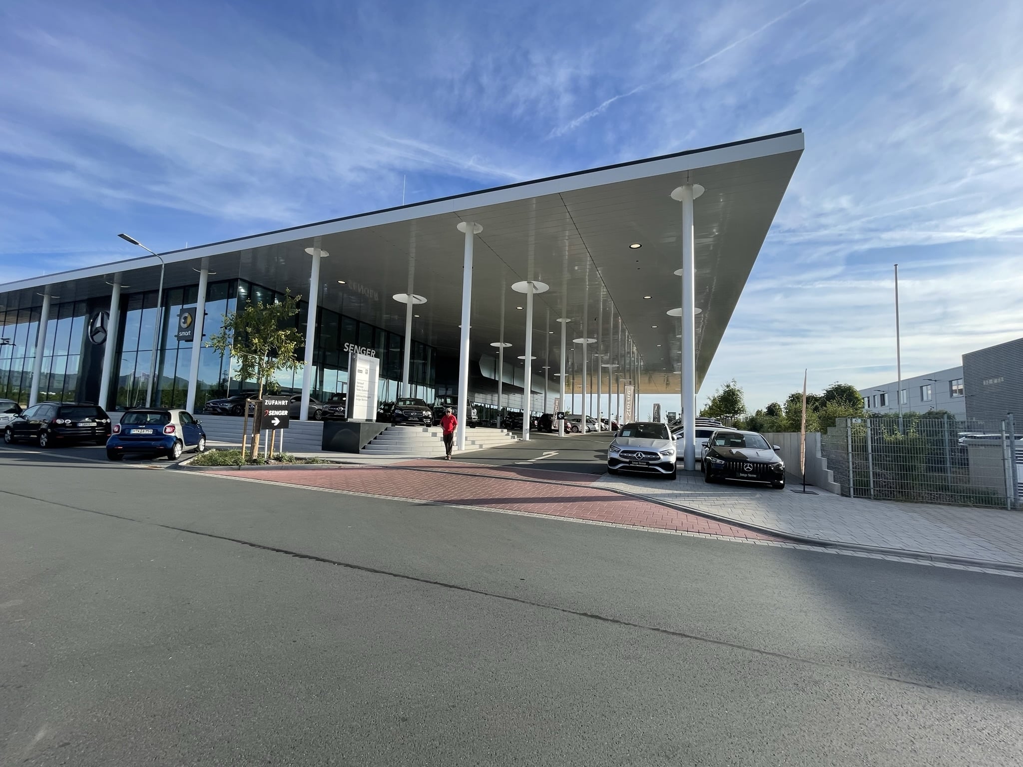 Weitwinkelfoto des modernen Mercedes-Benz-Autohauses in Oberursel mit auskragendem Dach, vielen weißen Stützen, großzügiger Glasfassade und mehreren ausgestellten Fahrzeugen im Außenbereich bei sonnigem Wetter.