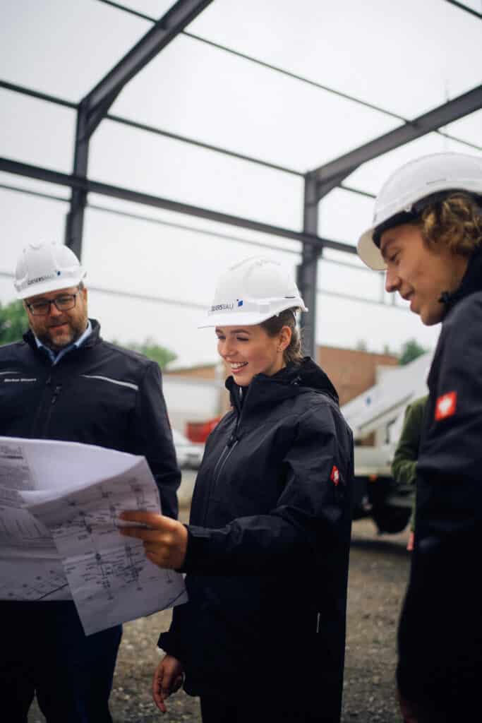 3 Personen mit Bauhelm schauen sich auf einer Baustelle einen Plan an.