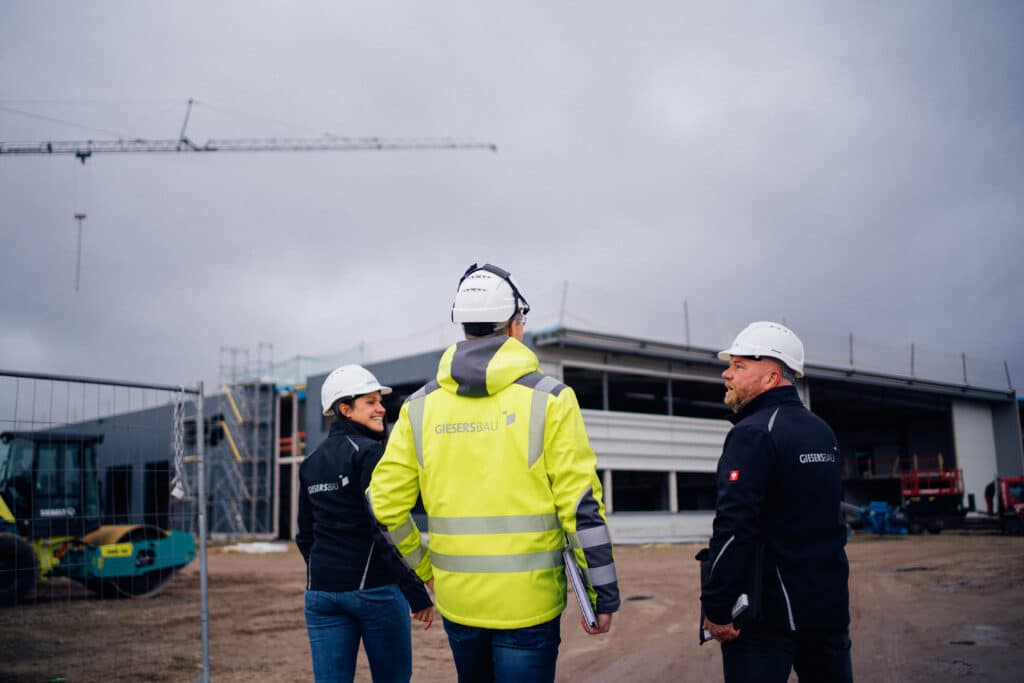 Drei Bauleiter:innen mit Schutzhelmen und Warnkleidung besprechen sich auf einer Baustelle vor einem Industriegebäude im Bau.