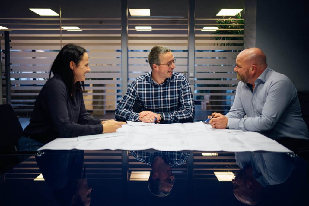Drei Personen sitzen an einem Konferenztisch und besprechen Baupläne; zwei Männer und eine Frau schauen einander zugewandt an.