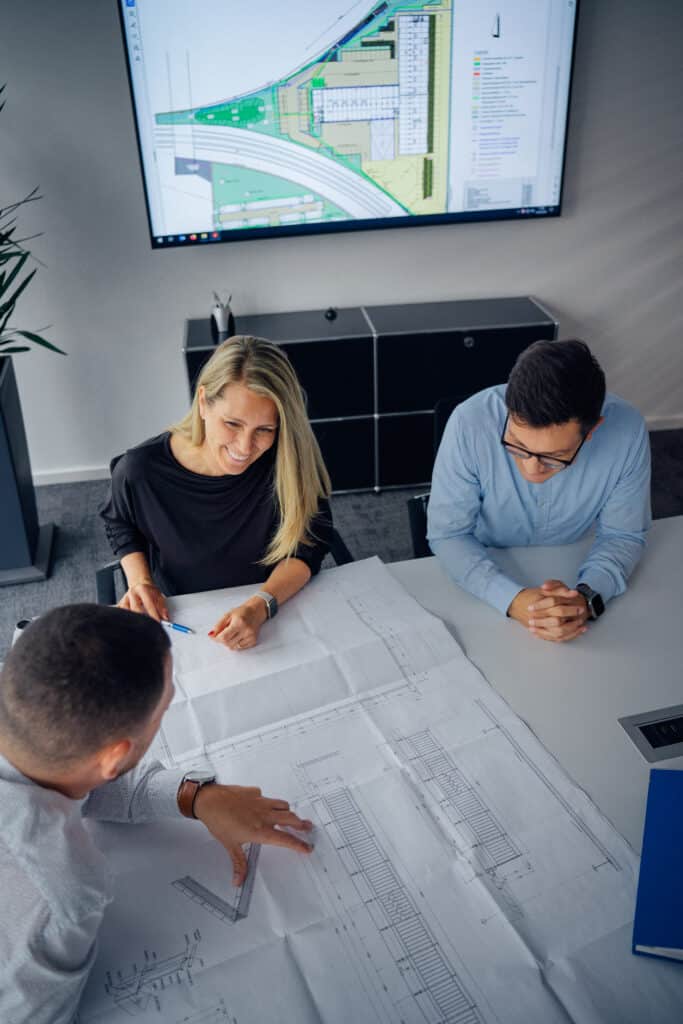 Drei Personen in einer Baubesprechung mit einem Konstruktionsplan vor sich auf dem Tisch.