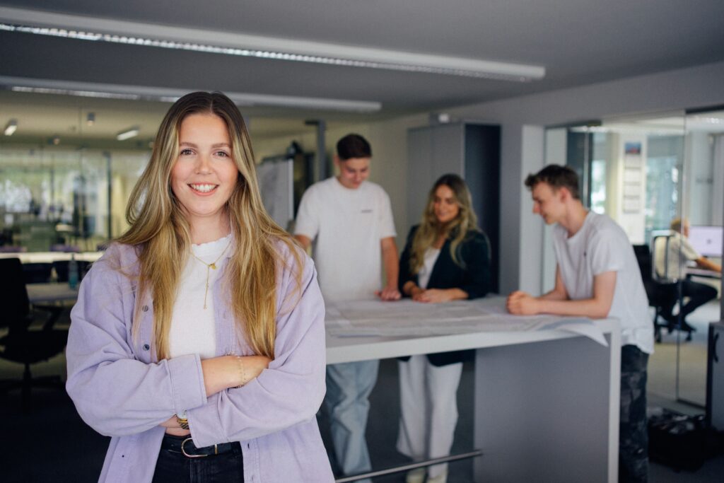 Mitarbeitende von Giesers Stahlbau GmbH um einen Tisch mit Bauplänen stehend. Im Vordergrund steht eine Mitarbeitende mit verschränkten Armen.