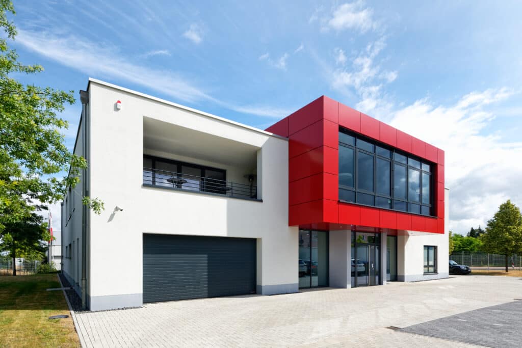 Modernes Büro- und Werkstattgebäude mit weißer Putzfassade, markant auskragendem roten Fassadenelement und großflächigen Glasfronten im Eingangsbereich.