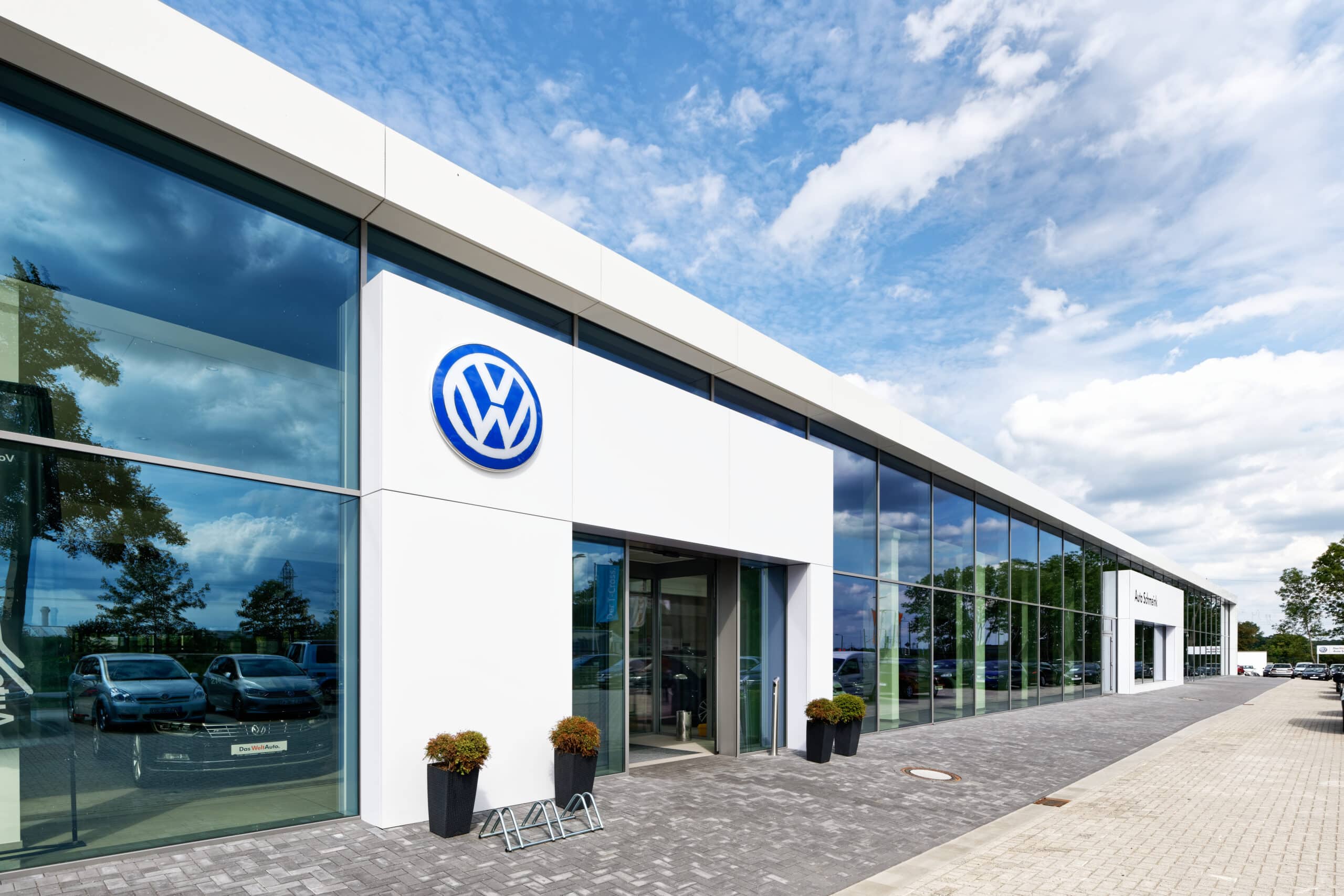 Moderne Außenansicht eines Autohauses in Wesel mit großflächiger Glasfassade, weißem Fassadenrahmen und prominentem VW-Logo über dem Eingangsbereich bei leicht bewölktem Himmel.