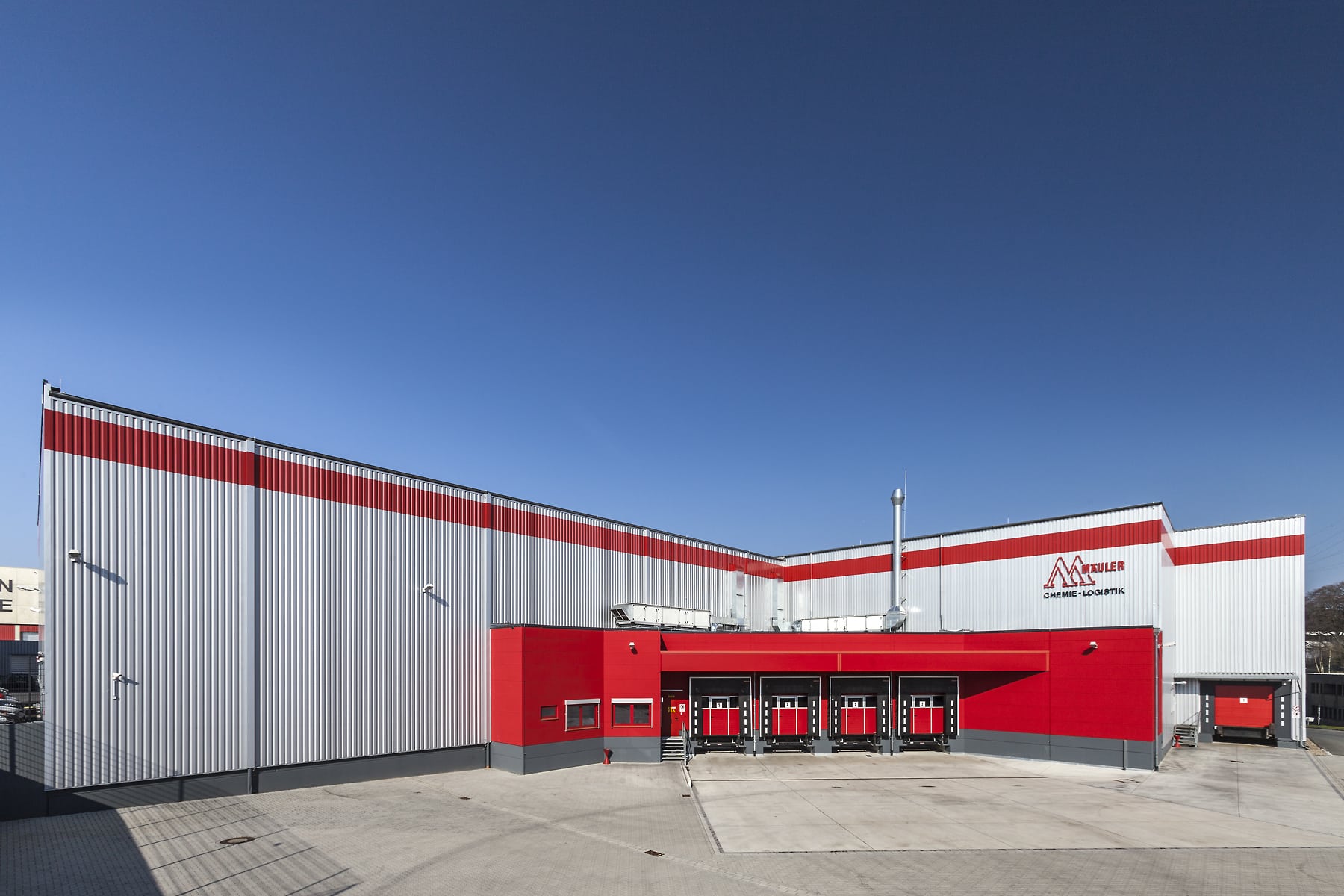 Große Lagerhalle in Remscheid mit markanter rot-silberner Fassade und mehreren überdachten Ladepunkten im Verladebereich. Im Vordergrund befindet sich eine weitläufige, gepflasterte Hoffläche.