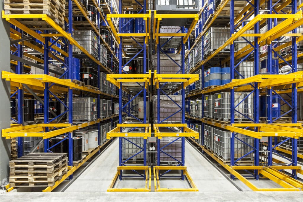 Innenansicht eines Hochregallagers mit farbigen Regalsystemen in Blau und Gelb. In den Regalen stehen große IBC-Behälter, Fässer und andere Industriegebinde, geordnet auf mehreren Ebenen entlang eines mittigen, schmalen Ganges.