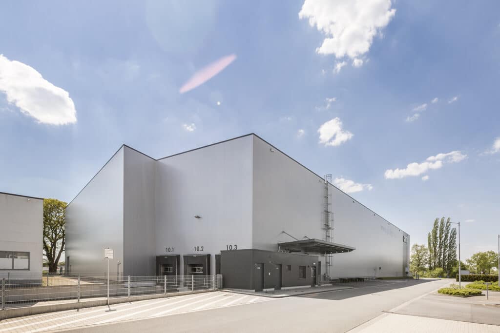 Seitenansicht des neu errichteten Hochregallagers in Bocholt mit hoher, weiß-grauer Sandwichpaneel-Fassade und klarer, kubischer Gebäudestruktur. Im Vordergrund befinden sich drei nummerierte Verladestationen (10.1–10.3) mit Andocktoren sowie ein vorgelagertes technisches Funktionsgebäude mit Vordach. Ein außenliegender Steigleiterzugang verläuft an der Hallenfassade. Die Anlage liegt an einer ruhigen Erschließungsstraße, umgeben von Grünflächen und Bäumen.