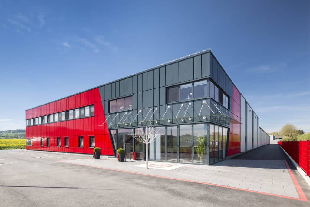 Neubau eines modernen Produktions- und Verwaltungsgebäudes in Neuenrade-Küntrop mit farblich akzentuierten Isowand-Paneelen in Rot und Anthrazit. Die zweigeschossige Fassade kombiniert großflächige Glasfronten im Eingangsbereich mit klar strukturierten Paneelflächen. Ein filigranes Glasvordach mit Edelstahl-Abhängungen betont den Haupteingang. Das Gebäude steht auf einem weitläufigen, befestigten Gelände, umgeben von offenen Landschaftsflächen.