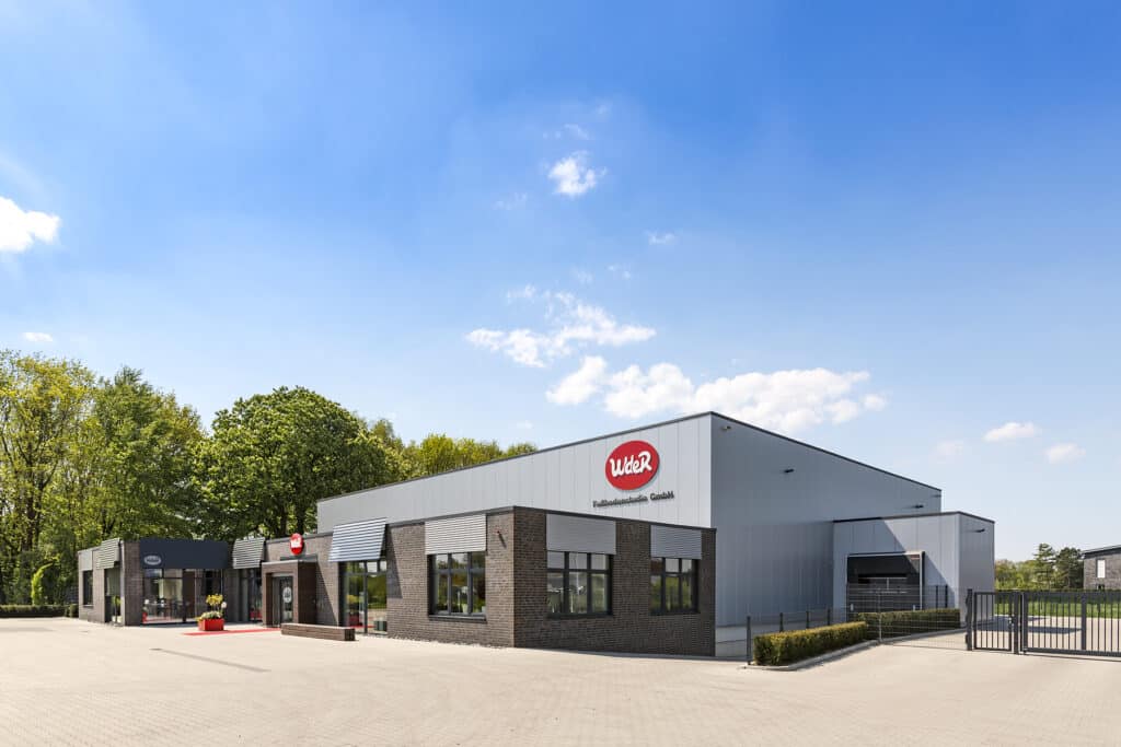 Moderner Betriebsneubau in Bocholt mit Kombination aus dunkelgrauem Klinkersockel und hellen Metall-Sandwichpaneelen. Das Gebäude verfügt über große Fensterflächen mit außenliegenden Lamellen, einen markanten Eingangsbereich und eine weitläufige, gepflasterte Hoffläche. Rechts befindet sich ein Zufahrtstor sowie ein angedocktes Hallentor mit Überladebereich. Im Hintergrund stehen Bäume, die das Grundstück einrahmen.