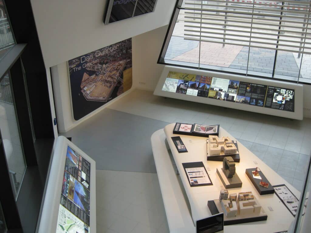 Innenraum des Infopavillons Überseequartier in Hamburg, fotografiert aus der oberen Etage. Zu sehen ist eine moderne Ausstellung mit Architekturmodellen auf weißen Podesten, großformatigen Informationstafeln an den Wänden und einer breiten Fensterfront mit horizontalen Lamellen, die Tageslicht in den Raum lässt.