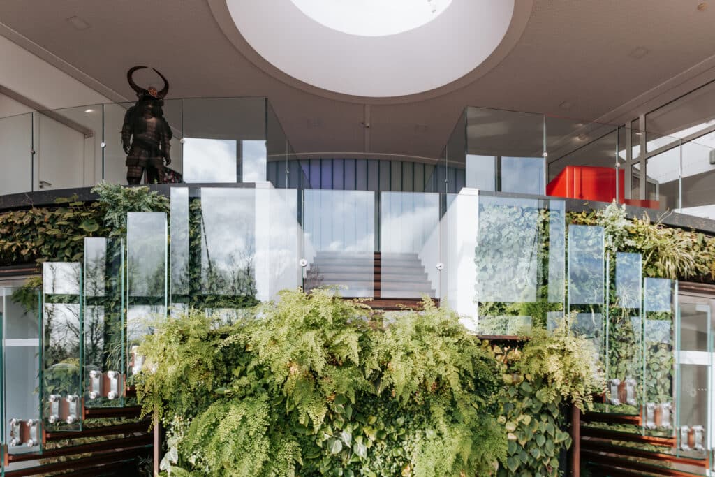Begrünter Innenraum mit Glasbrüstungen, zentraler Treppe, Oberlicht und einer Samurai-Rüstung auf der Galerie.