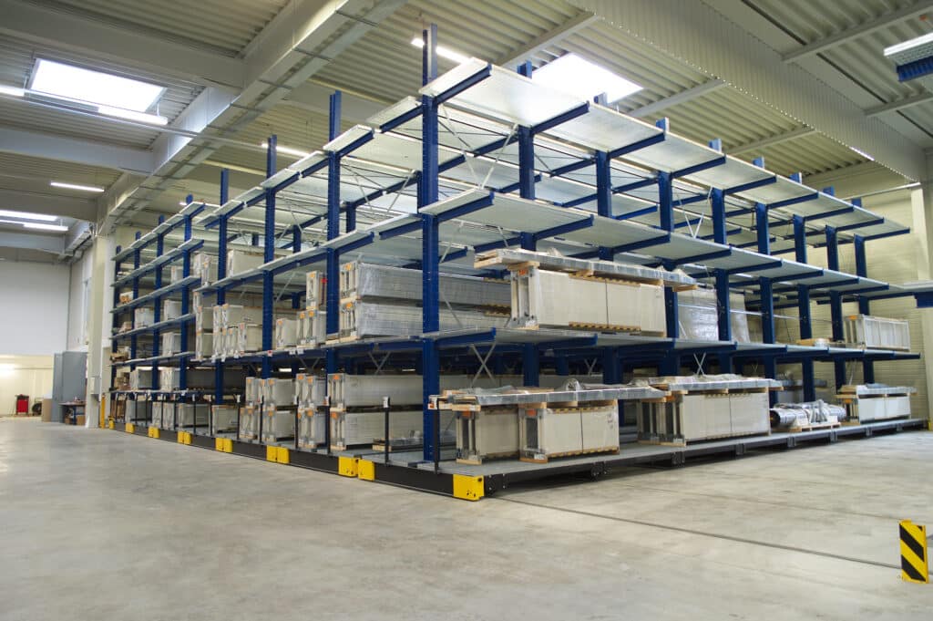 Innenansicht einer modernen Industriehalle mit einem hohen, blau lackierten Verschieberegal-System, auf dem verpackte Metall- und Bauelemente ordentlich eingelagert sind; die Halle ist hell beleuchtet und verfügt über ein graues Betonboden- und Stahlträgerdach.
