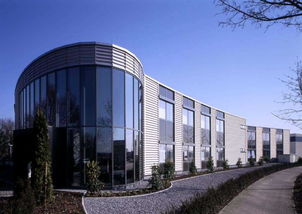 Moderner Neubau einer Produktions- und Lagerhalle mit vorgelagertem Sozialtrakt in Bocholt. Die geschwungene Glasfassade des Eingangsbereichs geht in eine strukturierte Metallfassade über. Große Fensterfronten, klarer Linienverlauf und eine gepflegte Außenanlage prägen die architektonische Gestaltung.